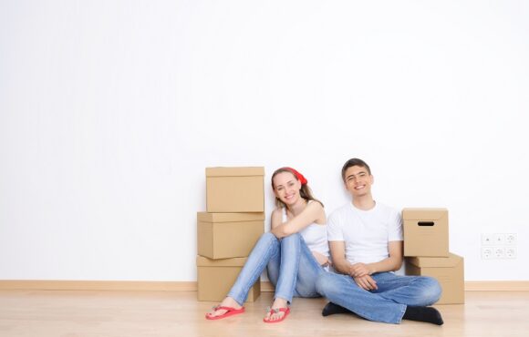 Młodzi kobieta i mężczyzna siedzą na podłodze pustego mieszkania, otoczeni kartonowymi pudełkami