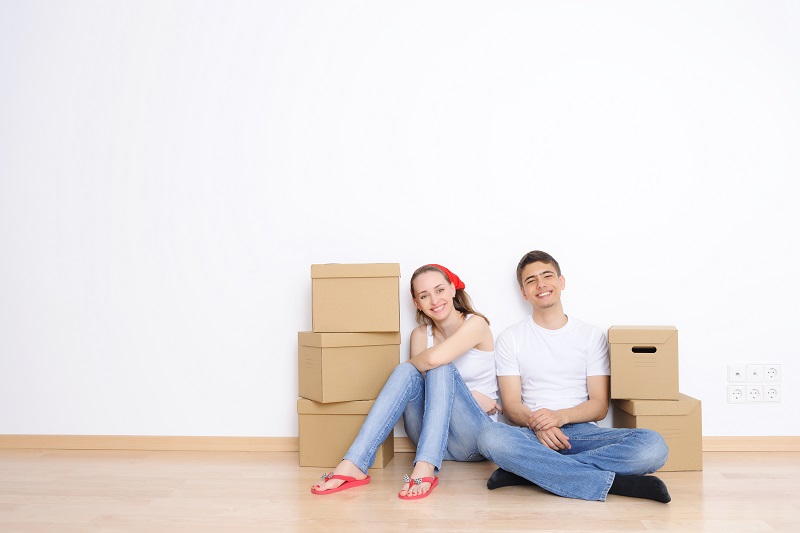 Młodzi kobieta i mężczyzna siedzą na podłodze pustego mieszkania, otoczeni kartonowymi pudełkami